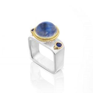 Sterling silver, square ring, with 10 mm moonstone set in 18ky gold and two .10 ct sapphires set in 18k yellow gold. Tapered band 9 mm to 7 mm wide x 1.5 mm thick.