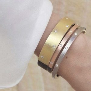 Three bangles on a wrist, one wide gold with inset scattered diamonds, and two thin in rose gold and white gold with inset diamonds spaced acros the top
